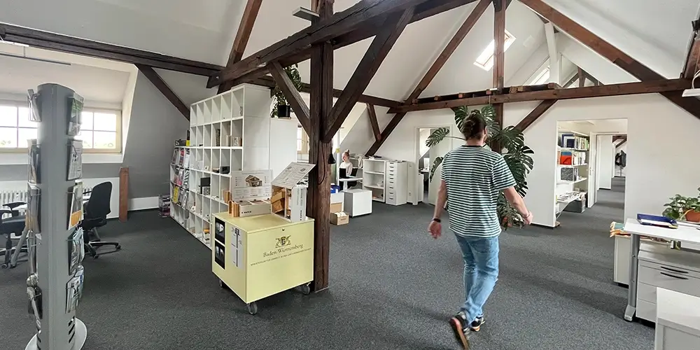 Modernes Großraumbüro mit offenen Holzbalken, Regalen und Arbeitsplätzen; Person geht durch den Raum