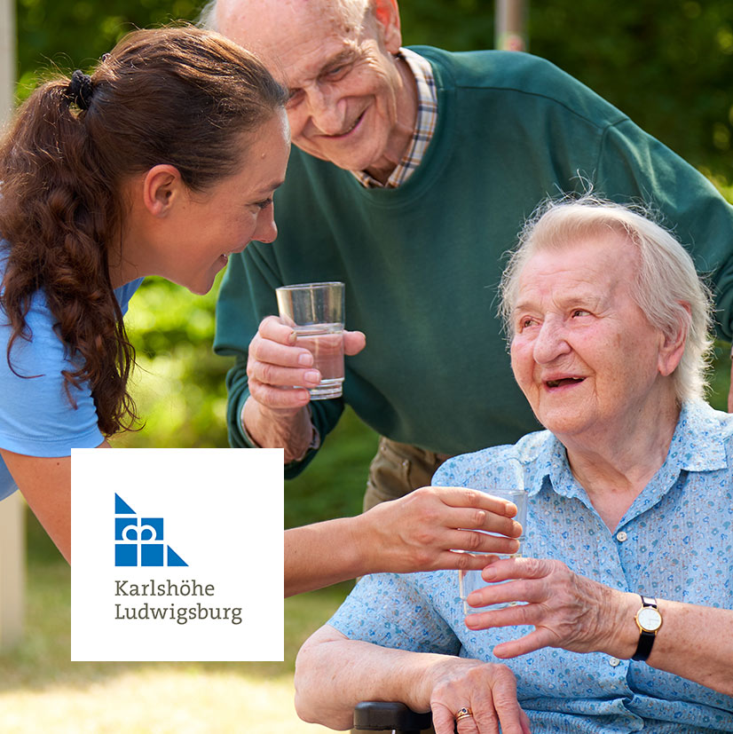 Eine Pflegekraft der Karlshöhe Ludwigsburg unterstützt zwei Senior*innen einfühlsam beim Trinken im Garten. Altenpflege, Betreuung und wertschätzende Unterstützung im Alltag.