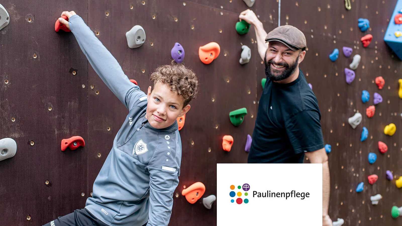 Paulinenpflege Bild, 2 Personen, die an einer Kletterwand hängen und in die Kamera lächeln.