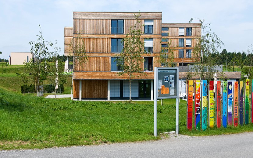 Fachklinik Höchsten | Modernes Holzgebäude mit bunten, bemalten Zaunpfählen im Vordergrund auf einer grünen Wiese